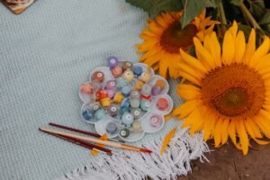 paint by number supplies, paintbrushes, and bright sunflowers arranged on a picnic blanket for a relaxing outdoor summer painting activity