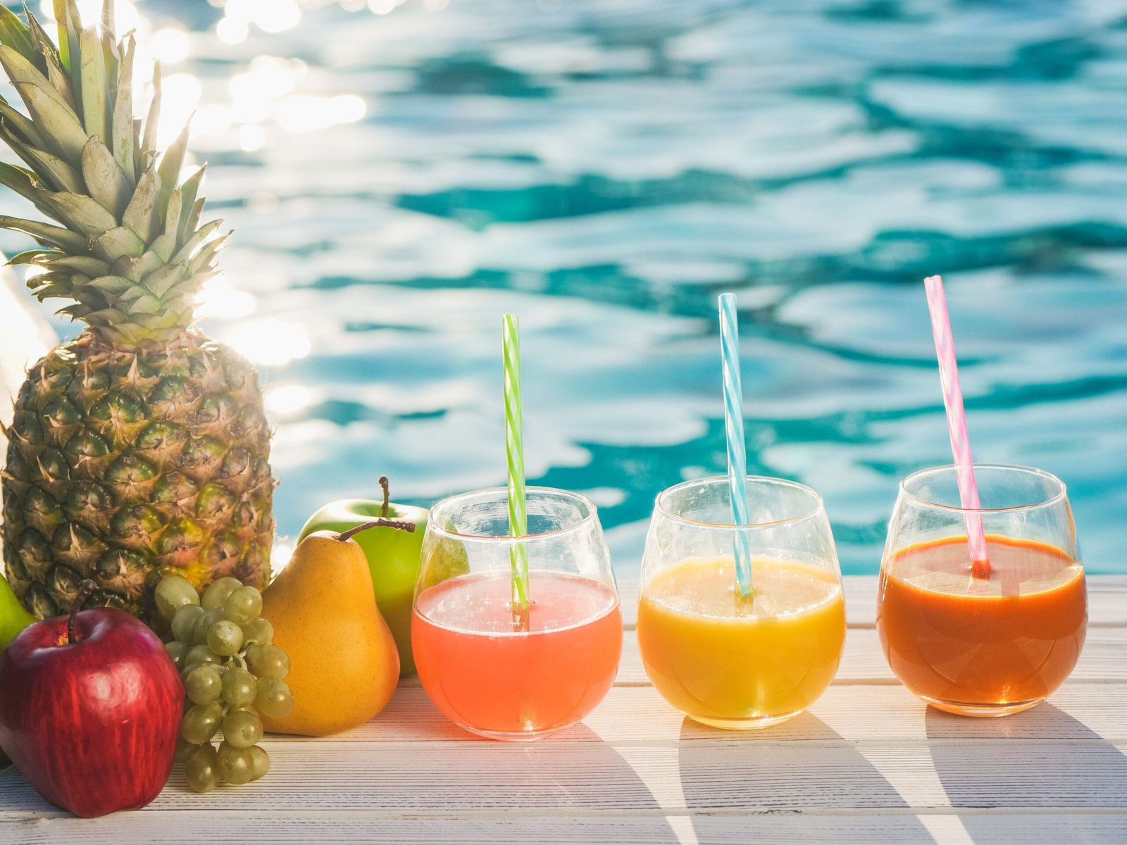 Fresh fruit and colorful fruit juices on a wooden table beside a sparkling outdoor swimming pool on a sunny summer day