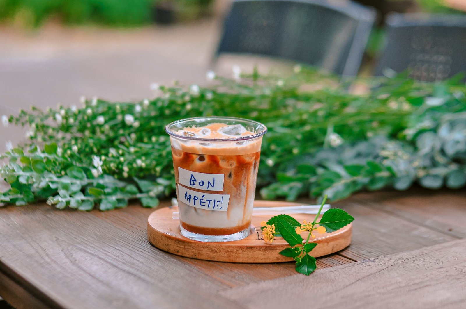 Glass of iced latte on a wooden table at an outdoor café surrounded by fresh greenery on a peaceful summer day.