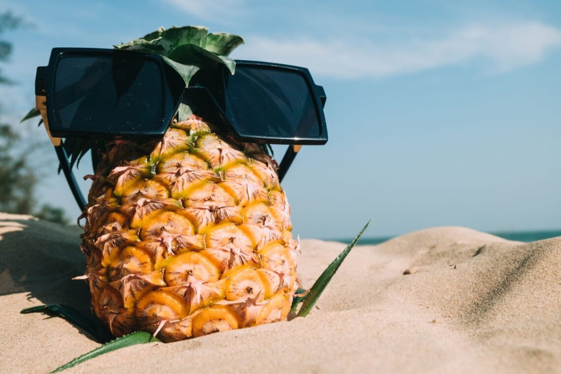 Pineapple wearing an oversized sunglasses on a sandy beach under a bright blue summer sky.