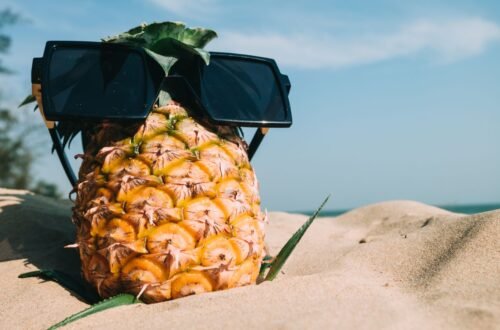 Pineapple wearing an oversized sunglasses on a sandy beach under a bright blue summer sky.