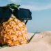 Pineapple wearing an oversized sunglasses on a sandy beach under a bright blue summer sky.