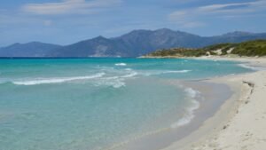 Turquoise waves washing into a white sandy beach with mountains in the distance under a clear blue summer sky.