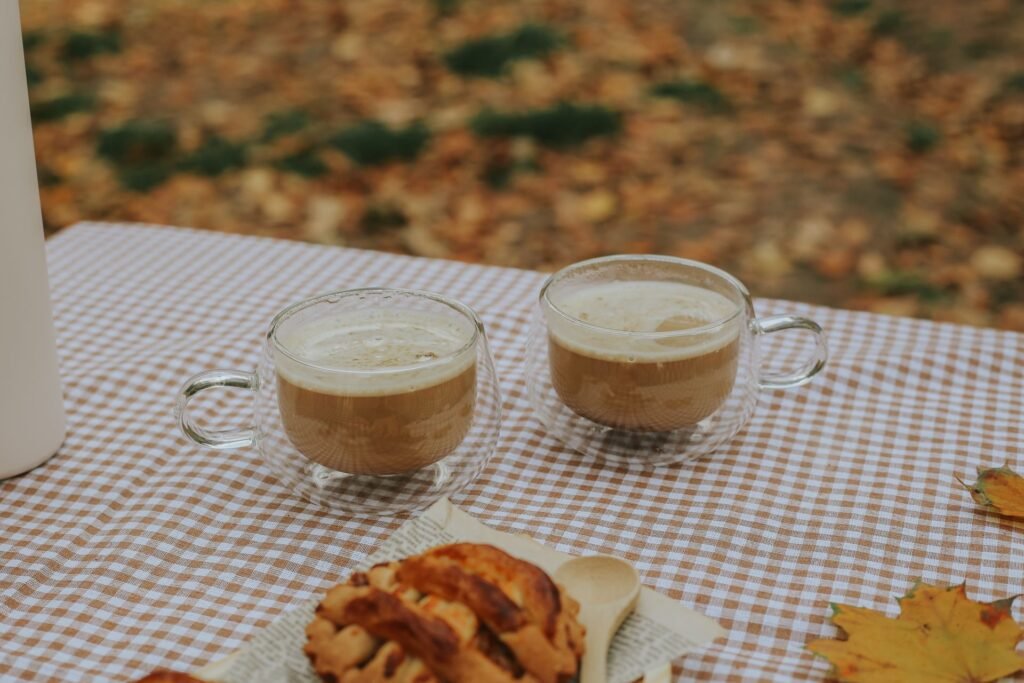 Two cups of coffee and a pastry on a checkered tablecloth outdoors, surrounded by autumn leaves