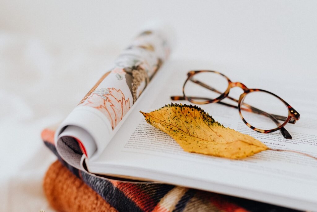 Open book with a yellow autumn leaf and glasses, resting on a cozy knitted blanket