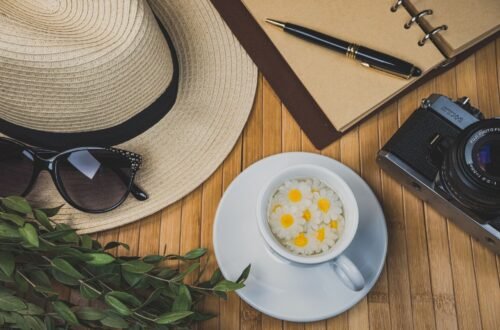 Summer flatlay with a camera, sunglasses, journal, flowers, and iced drink on a wooden table