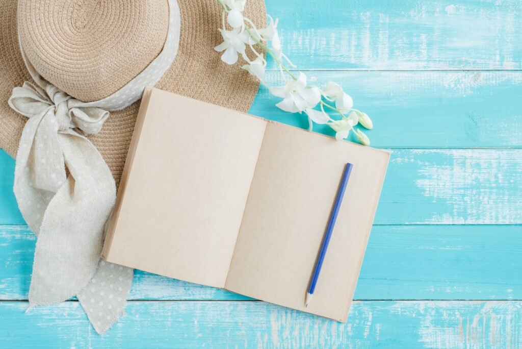 Summer flat lay with an open journal, blue pen, straw hat, and flowers on a turquoise table