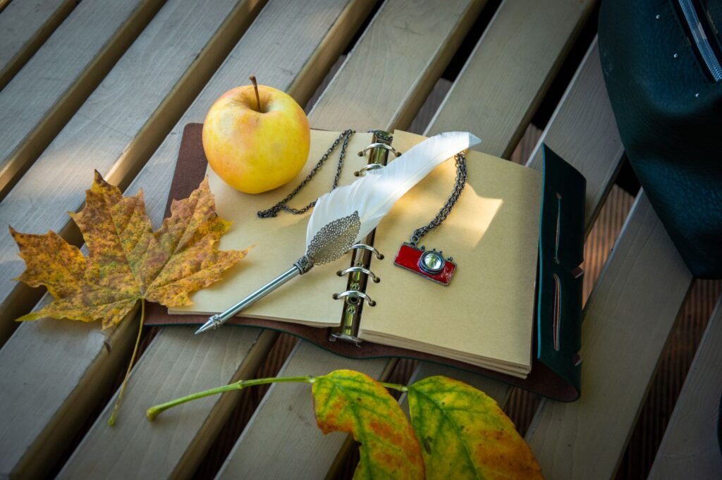 Open journal with a feather pen, apple, autumn leaves, and vintage camera keychain on a wooden bench
