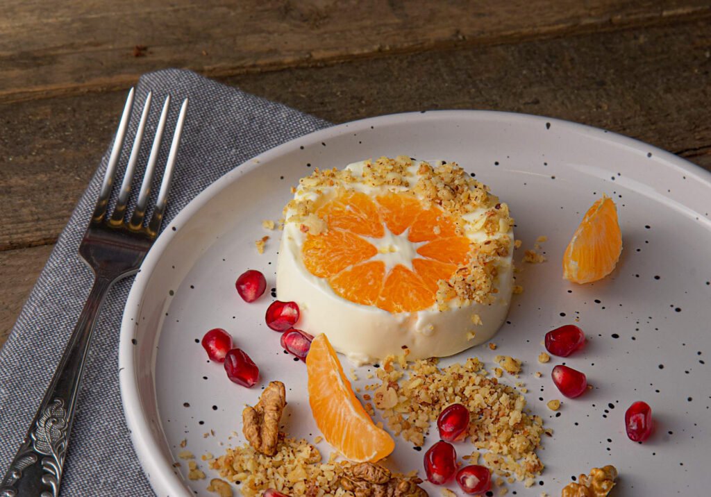 A small citrus dessert topped with orange slices and nut crumble, served on a white plate with pomegranate seeds and orange segments on a rustic wooden table.
