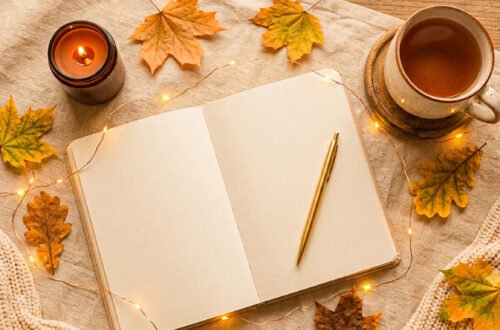 Cozy fall journaling setup with an open journal, gold pen, warm drink, candle, autumn leaves, and string lights on a neautral fabric surface