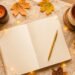 Cozy fall journaling setup with an open journal, gold pen, warm drink, candle, autumn leaves, and string lights on a neautral fabric surface