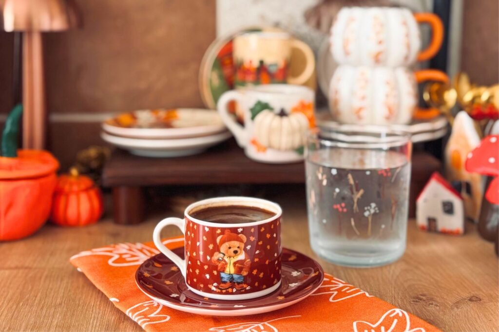 cozy autumn coffee setup with a red patterned mug on a wooden table, surrounded by pumpkins, seasonal mugs, and warm fall decor