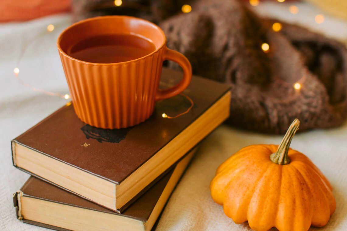 Cozy fall scene with an orange mug on a stack of books, a small pumpkin, a folded brown knit sweater, and warm string lights on a soft cream background.